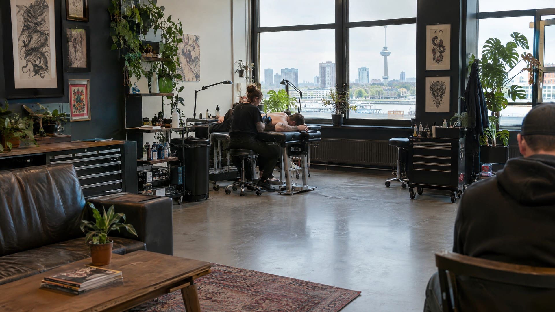 Tattoostudio met hoge stalen ramen, een tatoeëerder aan het werk en uitzicht over het water en de skyline van Rotterdam